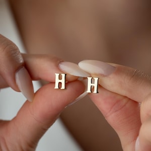May include: A pair of small, gold-toned stud earrings, each featuring the capital letter H in a classic serif font. The earrings have a polished, reflective surface and are held between fingers to display their size and shape clearly.