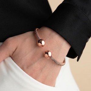 May include: A rose gold cuff bracelet featuring a textured, woven-style band that terminates in two polished, spherical ball ends. The bracelet is open-ended, allowing it to be worn around the wrist with the two spheres facing each other.