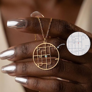 May include: A gold-colored necklace with a circular pendant featuring a map design and a small blue gemstone. The necklace is held up by a hand with silver nail polish, and a map of a city is shown in the background.
