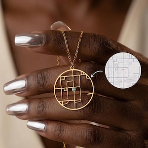 Könnte beinhalten: Eine goldfarbene Halskette mit einem kreisförmigen Anhänger mit einem Kartendesign und einem kleinen blauen Edelstein. Die Halskette wird von einer Person mit silbernem Nagellack gehalten. Eine kleine Karte ist im Hintergrund.