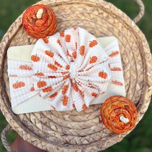 Könnte beinhalten: Ein weißes Stirnband aus Waffelstrick mit einem sich wiederholenden orangefarbenen Kürbismuster, das zu einem Knoten gerafft ist. Das Stirnband liegt auf einer rechteckigen Holzkarte, ergänzt durch zwei dekorative geflochtene Kürbisse.