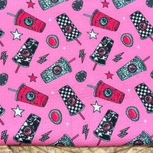 May include: A bright pink fabric featuring a repeating pattern of various drink cups with straws, interspersed with small white stars, black lightning bolts, and oval-shaped accents. The cups display diverse designs including checkered, cow print, and teal patterns.
