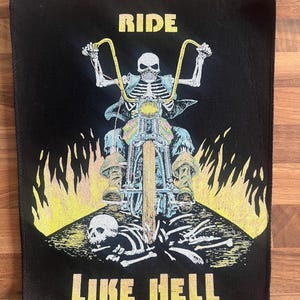 以下が含まれることがあります： 黒いパッチ。骸骨がバイクに乗っているデザインで、「RIDE」と「LIKE HELL」の文字が黄色で描かれています。炎と頭蓋骨も描かれており、バイカーをテーマにしたデザインです。