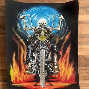 May include: Black graphic of a skeleton on a motorcycle, set against flames and a blue backdrop. The skeleton grips the handlebars, and the motorcycle has chrome detailing and a yellow headlight. The image evokes a biker theme.