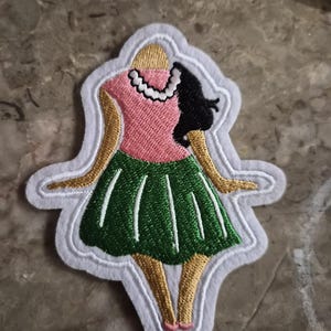 May include: An embroidered patch featuring a figure with tan skin, black hair, and a white necklace, wearing a pink top and a green skirt. The design is outlined in white thread and has a white fabric border, showcasing detailed stitching throughout.
