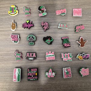 May include: A selection of colourful shoe charms with diverse designs and text. The charms feature pink, green, black, and white. Text includes "AKA", "1908", "Pretty Girl", "Pinkies", and "Danger Pretty Educated".