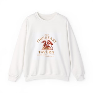 May include: A white sweatshirt featuring a logo for "The Fireheart Tavern." The logo includes a red dragon over an open book, with text that reads "Books, Brews & Community." The sweatshirt is a crew neck style.