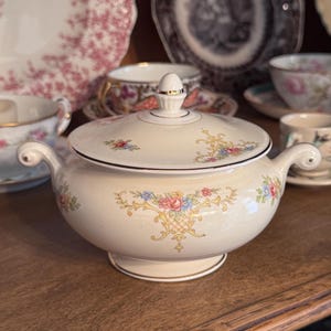 May include: A cream-colored porcelain sugar bowl with a matching lid, featuring delicate floral patterns in pink, blue, and yellow with gold filigree accents. The bowl has two curved handles and a small finial on the lid, all finished with gold trim.