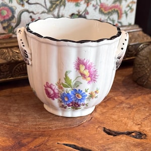 May include: A white ceramic sugar bowl with a fluted body and two handles. The exterior features a colorful floral pattern with pink, blue, and yellow flowers. The rim of the bowl is accented with a thin black line, and the handles have black detailing.
