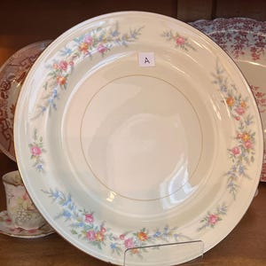 May include: A cream-colored dinner plate with a gold rim and floral design. The plate features a border of pink, blue, and orange flowers and foliage. The letter "A" is visible on the plate. The plate is displayed on a clear stand.