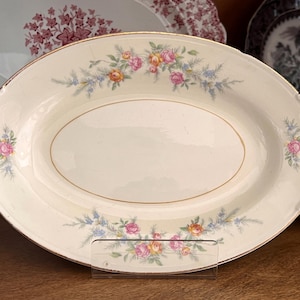 May include: Cream-colored oval serving platter with a gold rim and floral design. The platter features pink, yellow, and blue flowers and foliage. The edges have a scalloped design. Other decorative plates are in the background.