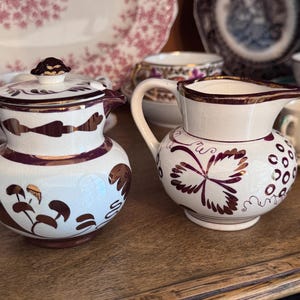May include: Two ceramic creamers with a white base and dark metallic brown floral and leaf patterns. The left creamer features a matching lid with a dark knob, while the right creamer has an open top with a pouring spout and a curved handle.