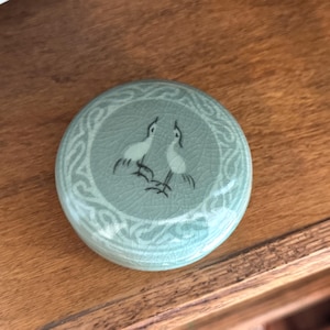 May include: A round, light green ceramic lid featuring a crackled glaze finish. The center displays a black line drawing of two birds facing each other, surrounded by a decorative border pattern of interlocking lines etched into the surface.