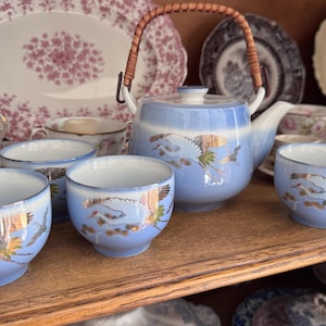 May include: A light blue ceramic tea set featuring a teapot with a woven handle and three matching tea cups. Each piece is decorated with a detailed illustration of a white crane in flight, accented with gold and green details against a blue gradient.