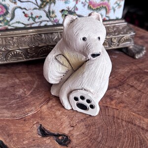 May include: A cream-colored bear figurine with a textured surface, featuring dark eyes, a dark nose, and a paw pad with five distinct dark circular accents. The bear is depicted in a seated position with one paw raised toward its face.