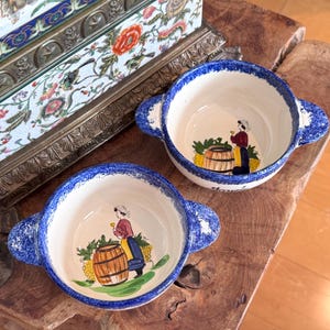May include: Two ceramic bowls with handles, each featuring a blue sponge-painted rim and a hand-painted illustration inside. The design depicts a person in a red top and blue skirt standing next to a wooden barrel with yellow grapes nearby.