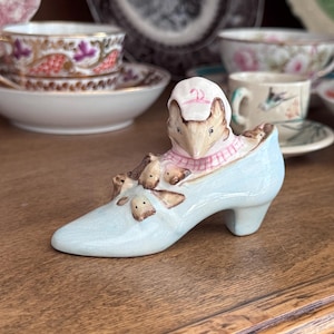 May include: A ceramic figurine of a fox wearing a pink and white checkered bonnet, nestled inside a light blue high-heeled shoe. The fox has brown fur and is surrounded by several small brown mice figures resting on the surface of the shoe.