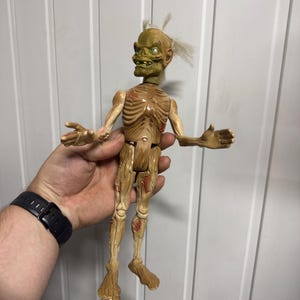 Pode incluir: Uma figura de ação articulada em tons de bege e verde de uma criatura esquelética com postura curvada, caixa torácica exposta e cabelo claro ralo. A figura apresenta texturas de pele moldadas detalhadas, articulações visíveis nos ombros, quadris e joelhos, e uma expressão facial ameaçadora com dentes proeminentes e olhos grandes.