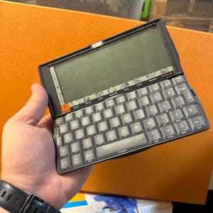 Könnte beinhalten: Ein dunkelgrauer Psion Series 5 Handheld-Computer mit QWERTY-Tastatur und aufklappbarem Bildschirm. Das Gerät verfügt über eine Reihe von Anwendungssymbolen wie System, Word, Sheet, Data, Agenda, Time, Calc und Sketch am unteren Displayrand.