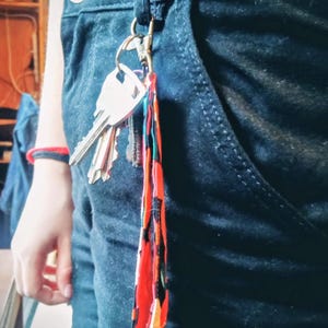 Peut inclure: Un trousseau de clés attaché à une lanière colorée, accrochée à une poche de jean bleu foncé. La lanière présente un motif vibrant de formes géométriques rouges, oranges, noires et vertes. Les clés sont argentées.