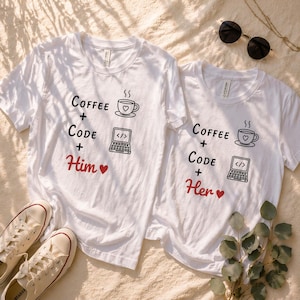 May include: Two white t-shirts with the text "Coffee + Code + Him" and "Coffee + Code + Her" printed in black, with a red heart. Each shirt features a coffee cup and a laptop graphic. A pair of white sneakers and sunglasses are also visible.