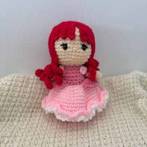 K&ouml;nnte beinhalten: Eine handgefertigte H&auml;kelpuppe mit roten Haaren und einem rosa Kleid. Die Puppe hat ein beiges Gesicht mit schwarzen Augen und tr&auml;gt ein zweifarbiges rosa Kleid mit wei&szlig;er Bord&uuml;re. Die Puppe sitzt auf einer cremefarbenen, strukturierten Oberfl&auml;che.
