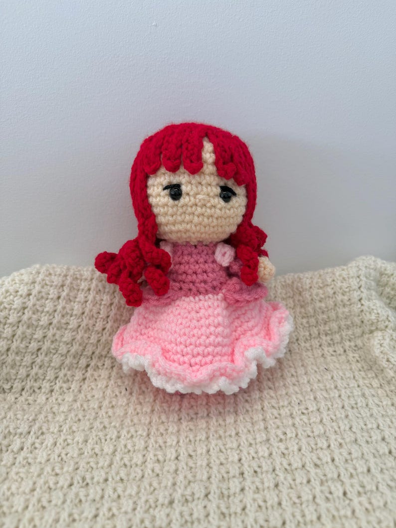 K&ouml;nnte beinhalten: Eine handgefertigte H&auml;kelpuppe mit roten Haaren und einem rosa Kleid. Die Puppe hat ein beiges Gesicht mit schwarzen Augen und tr&auml;gt ein zweifarbiges rosa Kleid mit wei&szlig;er Bord&uuml;re. Die Puppe sitzt auf einer cremefarbenen, strukturierten Oberfl&auml;che.