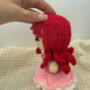 K&ouml;nnte beinhalten: Eine handgefertigte H&auml;kelpuppe mit roten Haaren und einem rosa Kleid. Das Haar der Puppe ist lockig gestylt und das Kleid hat eine wei&szlig;e Bord&uuml;re. Die Puppe wird von einer Hand mit einem goldenen Ring gehalten.