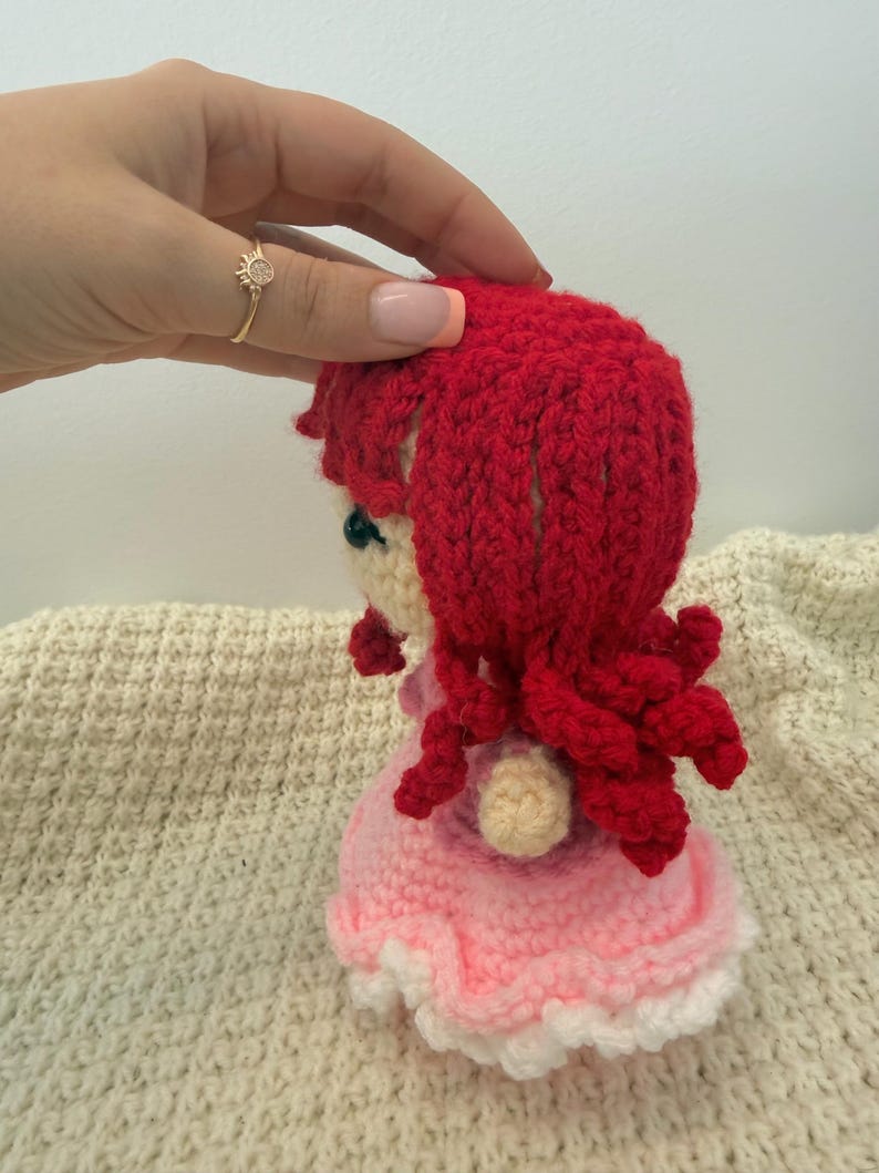 K&ouml;nnte beinhalten: Eine handgefertigte H&auml;kelpuppe mit roten Haaren und einem rosa Kleid. Das Haar der Puppe ist lockig gestylt und das Kleid hat eine wei&szlig;e Bord&uuml;re. Die Puppe wird von einer Hand mit einem goldenen Ring gehalten.