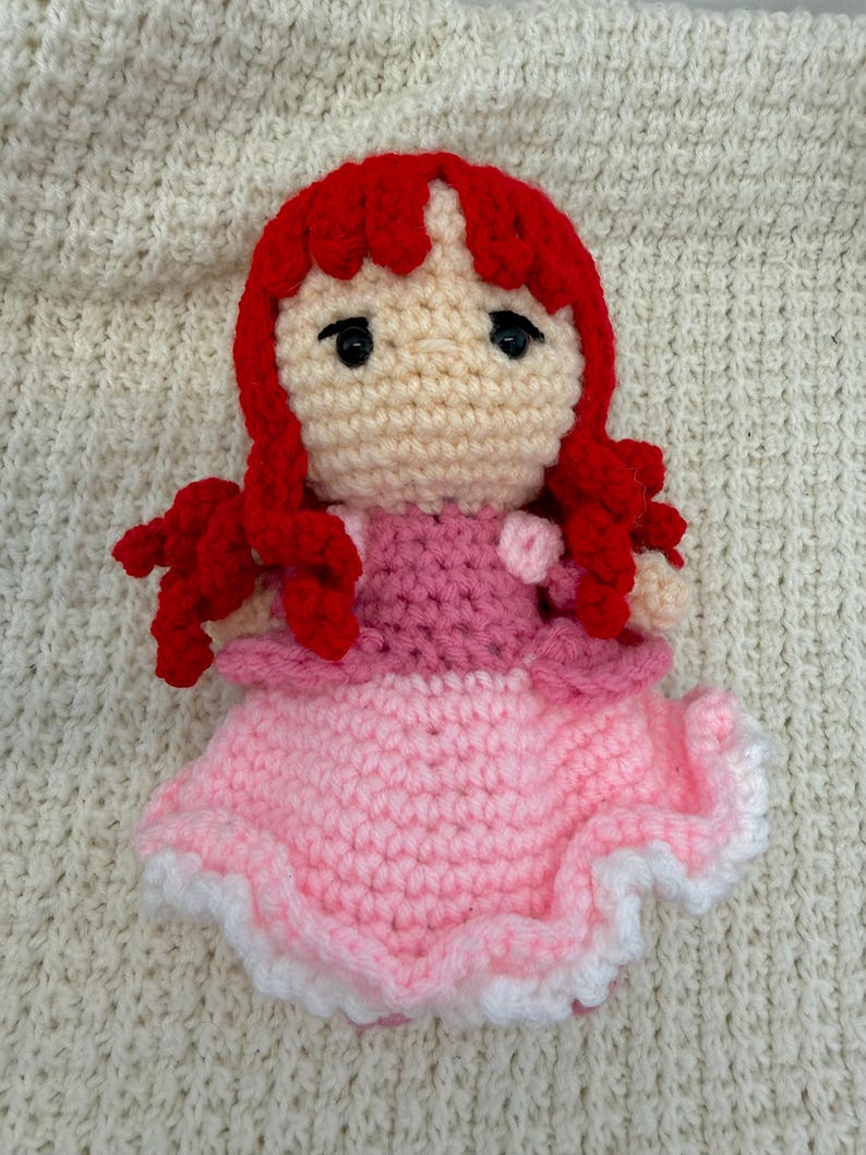 K&ouml;nnte beinhalten: Handgemachte H&auml;kelpuppe mit roten Haaren, schwarzen Augen und hellem Hautton. Die Puppe tr&auml;gt ein rosa Kleid mit wei&szlig;em Besatz. Die Puppe ist aus Garn gefertigt und sitzt auf einer cremefarbenen, strukturierten Oberfl&auml;che.