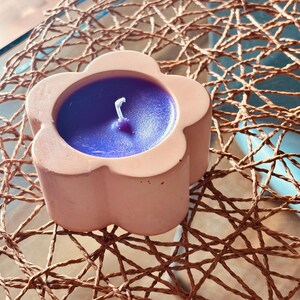 Peut inclure: Un bougeoir en forme de fleur fabriqué dans un matériau lisse de couleur pêche clair, contenant une bougie en cire violette vive avec une mèche en coton blanc centrée. Le récipient présente des pétales arrondis formant une forme circulaire.