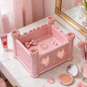 May include: A pink castle-shaped jewelry tray with heart-shaped cutouts and decorative turrets. The tray holds a pink bow, a gold ring, and pearl earrings. Makeup brushes, a compact, and a tube of lip gloss are also visible.