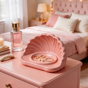 May include: A pink seashell-shaped jewelry tray holds rings and a bracelet. A perfume bottle, a candle in a glass, and a pearl hair clip sit nearby on a pink dresser. The bedroom has a pink bed with a tufted headboard.