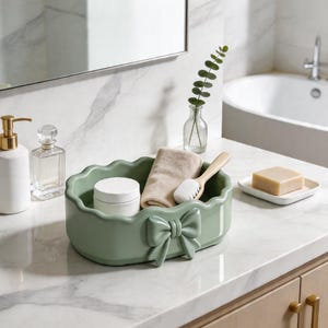 May include: A sage green ceramic bathroom organizer with a bow detail, holding a small towel, brush, and a jar of cream. Other bathroom accessories include a soap dispenser, perfume bottle, and a bar of soap.