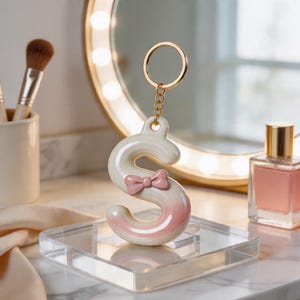 May include: A white and pink letter "S" keychain with a pink bow, displayed on a clear acrylic stand. The keychain has a gold-colored ring and chain. The background includes a vanity mirror and makeup brushes.