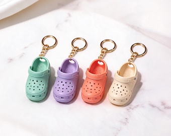 Puede incluir: Cuatro llaveros en miniatura con forma de zueco en colores pastel verde menta, lavanda, coral y crema. Cada llavero cuenta con una anilla de metal dorado unida por una cadena corta al talón de la réplica de zapato perforado.