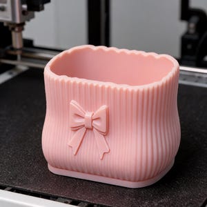 May include: A light pink, rectangular storage container with rounded corners and a vertical ribbed texture. The front of the container features a centred, moulded bow detail. The top edge has a scalloped design, and the base is flat.