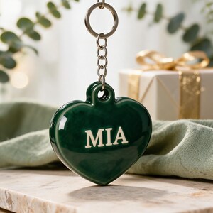 May include: A dark green heart-shaped keychain made of a smooth, glossy material. The center of the heart features the name MIA in bold, light-colored capital letters. The heart is attached to a silver-toned metal split ring by a short chain.