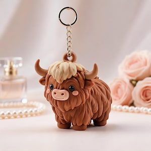 May include: A brown Highland cow keychain featuring a shaggy coat, a lighter tan tuft of hair on its head, small horns, and a pinkish snout. The figure is attached to a gold-toned metal chain and a circular ring for easy attachment to keys.