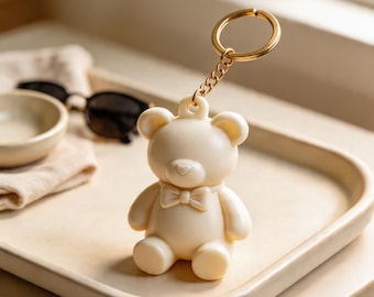 Puede incluir: Un llavero con forma de oso de color crema con un acabado liso y mate, una pequeña pajarita esculpida en el cuello y una anilla de metal dorado unida por una cadena corta. El oso tiene orejas redondeadas y una estructura facial sencilla.