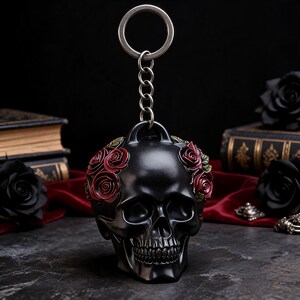May include: A black skull keychain featuring a crown of sculpted red roses with green leaves. The skull has a matte finish and is attached to a silver-toned metal split ring by a short chain, creating a detailed accessory for keys or bags.