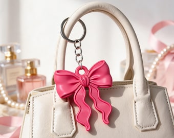 Puede incluir: Un llavero de lazo rosa brillante en tres dimensiones unido a una anilla de metal plateado. El lazo presenta largas cintas onduladas y cuelga del asa de un bolso de color crema, mostrando un acabado liso y de textura mate.