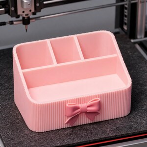 Puede incluir: Un organizador de escritorio de plástico rosa claro con una textura exterior acanalada y un lazo decorativo centrado en el panel frontal. El organizador incluye tres compartimentos superiores pequeños y un compartimento inferior más ancho.