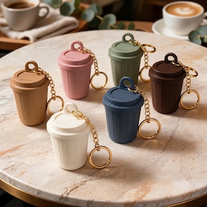 May include: A selection of miniature bin-shaped keyrings in shades of brown, pink, green, blue, white, and dark brown. Each keyring features a gold-coloured chain and ring. The keyrings are displayed on a marble-effect surface.
