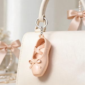 Op de afbeelding: Een lichtroze balletschoen-sleutelhanger met een kleine strik op de neus en gekruiste bandjes over de bovenkant. De miniatuurschoen is met een kort kettinkje bevestigd aan een zilverkleurige metalen ring en hangt aan een bijpassende handtas.