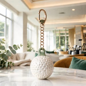 Puede incluir: Un llavero con forma de pelota de golf blanca con una superficie texturizada de hoyuelos y un lazo superior en color verde salvia. El llavero cuenta con una cadena de metal dorado y un mosquetón a juego para sujetar las llaves de forma segura.