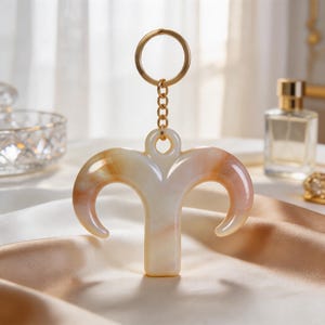 May include: A cream and orange Aries zodiac sign keychain with a gold-colored keyring and chain. The keychain is made of a translucent material and has a curved, horn-like design. The background includes a perfume bottle and a decorative dish.