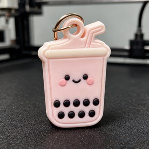 May include: A light pink bubble tea keychain featuring a smiling face with rosy cheeks and black boba pearls at the bottom. The cup has a cream-colored lid and a straw, with a gold-toned metal ring attached to the top for hanging purposes.