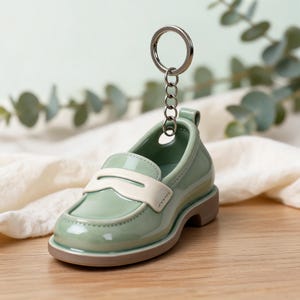 May include: A glossy sage green loafer keychain featuring a smooth finish, a cream-colored saddle strap across the vamp, and a sturdy silver-toned metal split ring attached to the heel collar for secure key storage and organization.