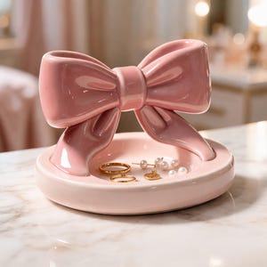 Puede incluir: Un plato para joyas de cerámica en color rosa claro brillante con un gran lazo esculpido en la parte posterior. La base circular contiene dos anillos dorados, un pequeño colgante de oro y varias cuentas blancas en el centro.